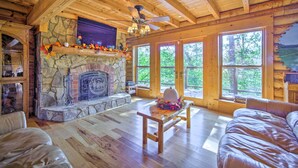 A cozy cabin interior with a stone fireplace, wooden beams, large windows, and a leather sofa. - Calhoun Cabin w/ River View + Wraparound Deck (Calhoun)