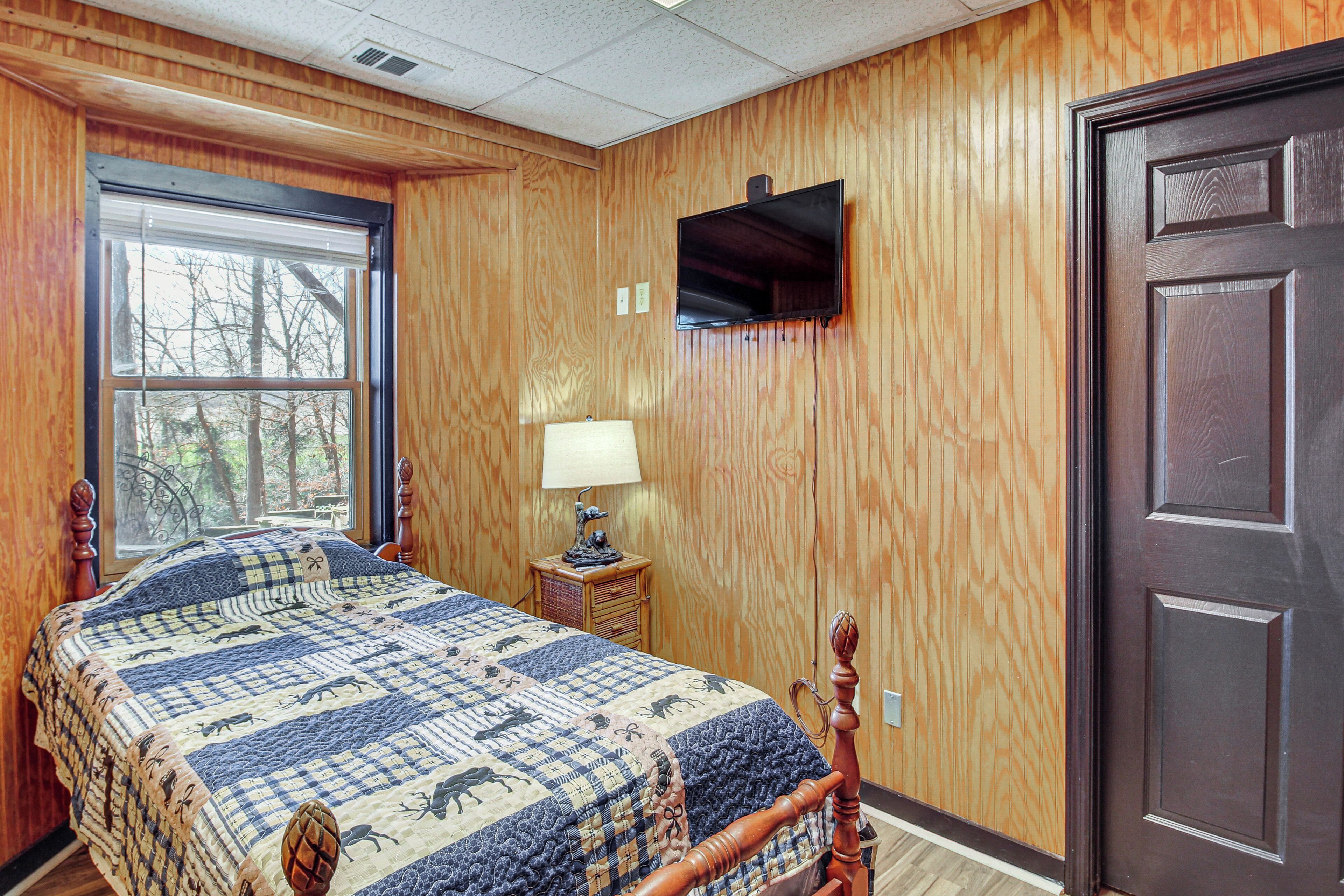 A bedroom with a wooden paneled wall, a bed with a quilt, a nightstand with a lamp, and a television mounted above a door.
