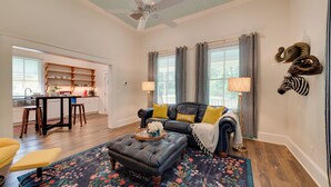A living room with a dark leather sofa, a wooden coffee table, and a floral rug. There are two windows with curtains, a ceiling fan, and a kitchen area in the background. - Restored Home Near Downtown Thomasville (Thomasville)