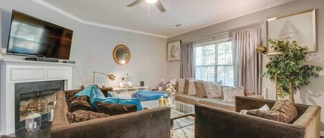 A modern living room with a fireplace, comfortable seating, a ceiling fan, and a large window with curtains.