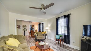 A living room with a sofa, coffee table, and a television. There is a ceiling fan, a lamp, and a window with curtains. - Charming Newport News Cottage < 1 Mi to Ocean! (Newport News)