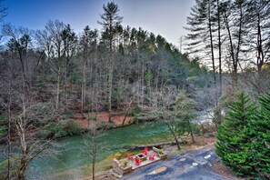 A riverside outdoor seating area with stone tables and red chairs, surrounded by dense forest and a clear river. - Riverside Ellijay Cabin w/ Hot Tub & Game Room (Ellijay)