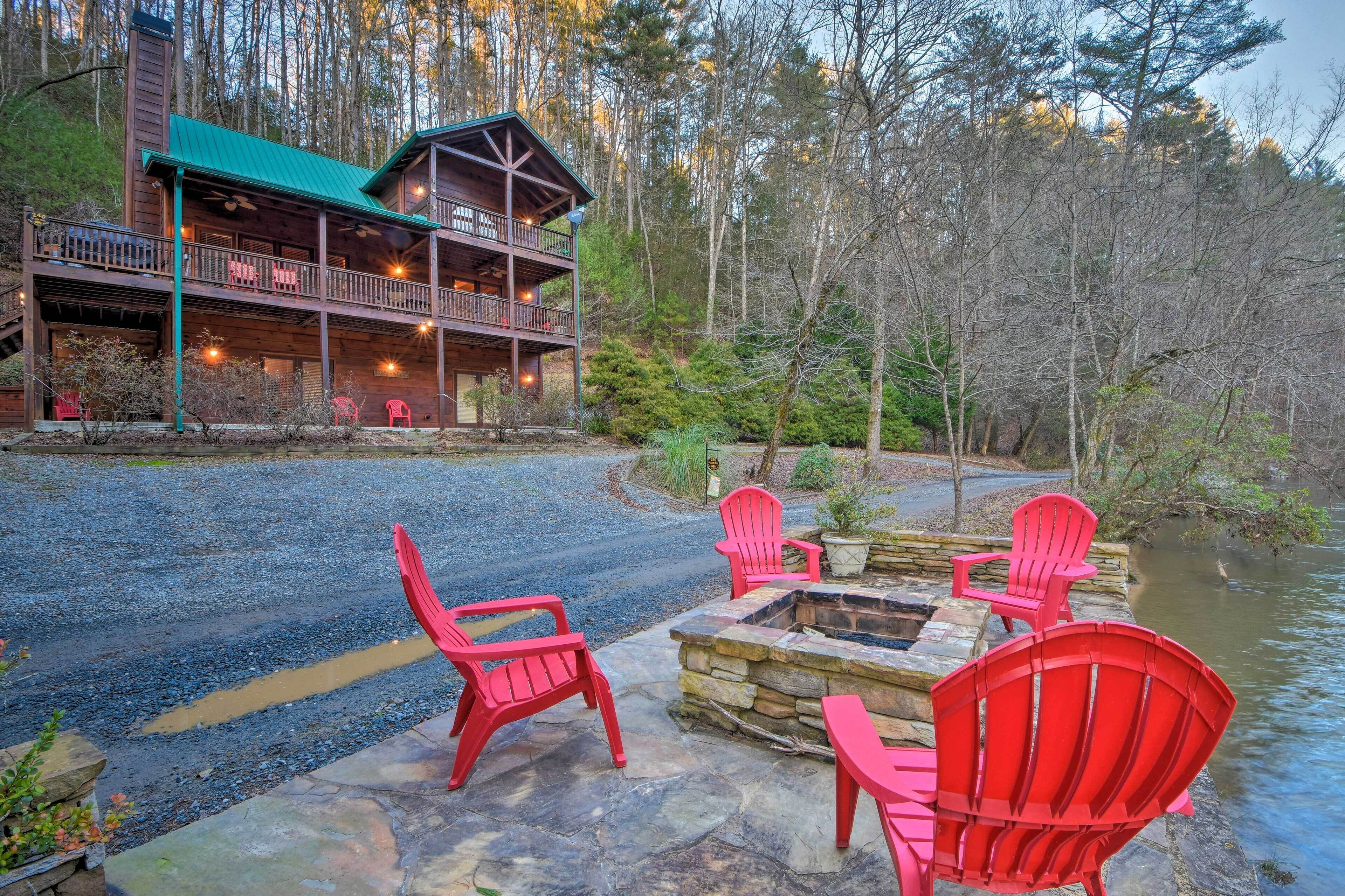 A lodge with a stone fire pit and red chairs, surrounded by a forest and a body of water.