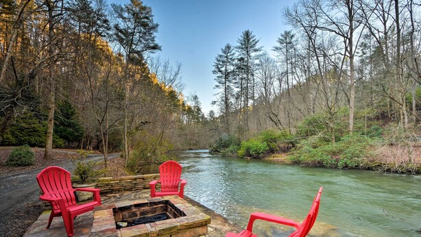 Un foyer en pierre, entouré de chaises Adirondack rouges, situé dans une zone boisée au bord d’une rivière.