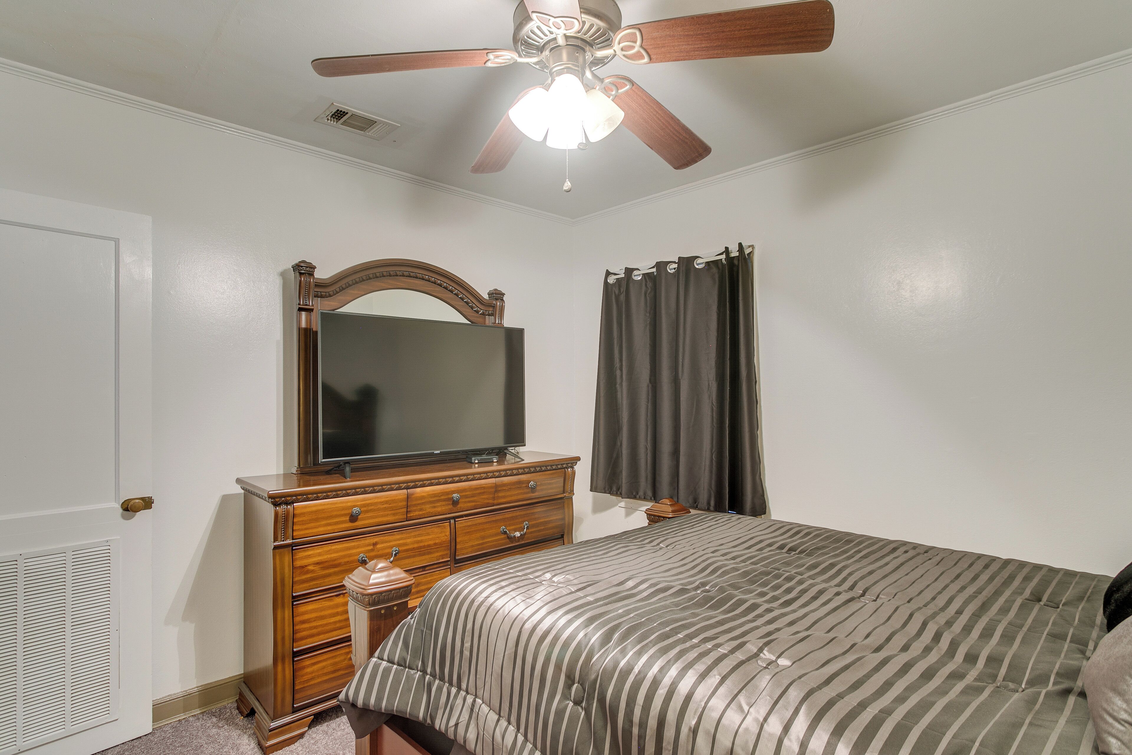 Une chambre à coucher comprenant un lit, une commode, un miroir, un ventilateur de plafond et une fenêtre avec des rideaux.