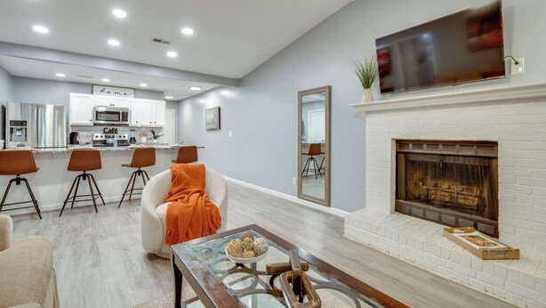 A modern living room with a fireplace, a dining area with bar stools, and a kitchen in the background.