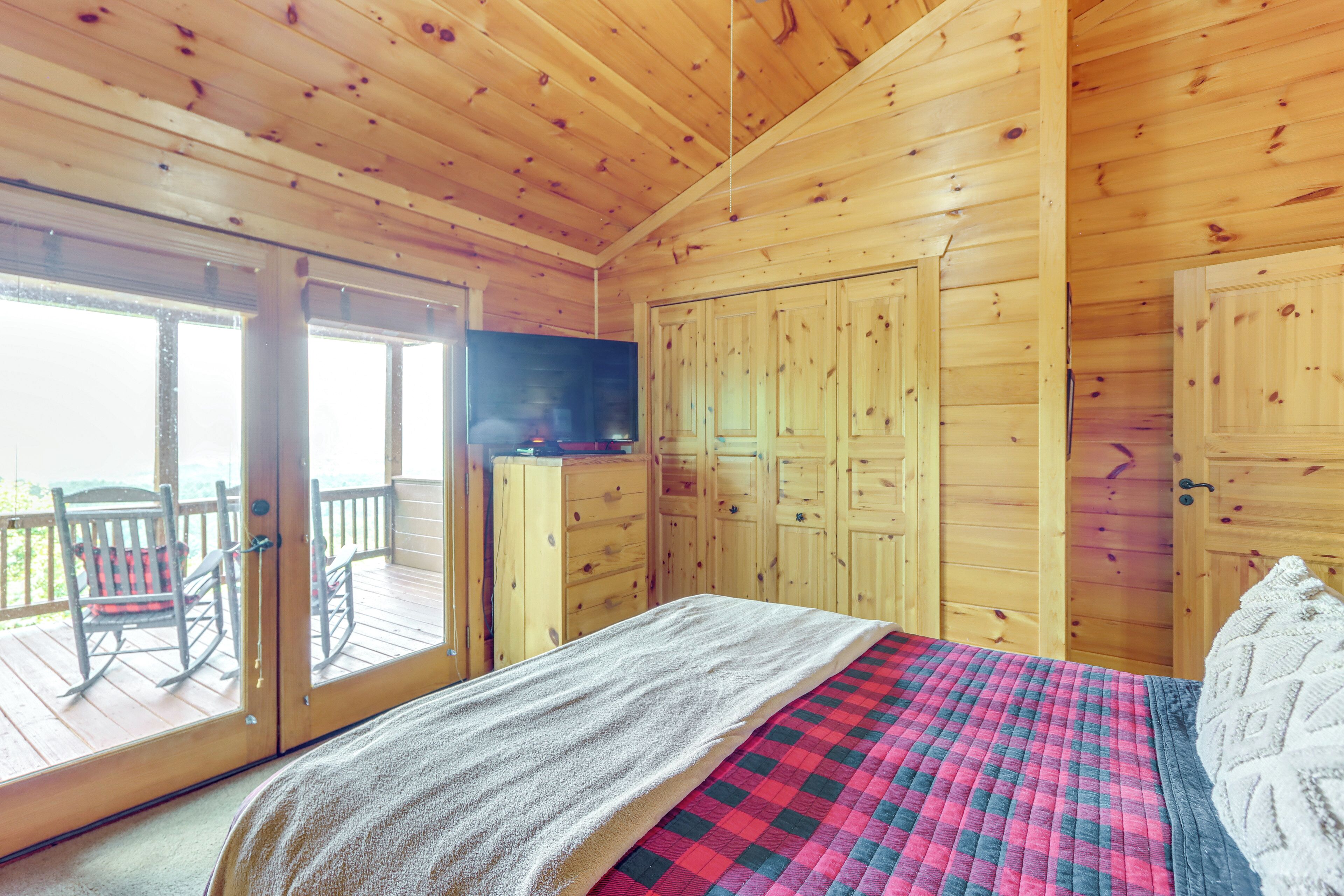 A wooden cabin bedroom with a bed, a TV, and a sliding glass door leading to a balcony with a chair.