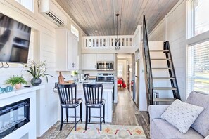 A compact kitchen with white cabinetry, a central island, and a microwave. There is a fireplace below the kitchen, a sofa with a patterned cushion, and a ladder against the wall. - Tiny Home Retreat w/ Fire Pit ~ 3 Mi to Lake! (Morganton)