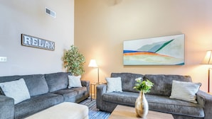 A living room with two grey sofas, a beige ottoman, a wooden coffee table, a floor lamp, a potted plant, and a wall-mounted abstract painting. - Horn Lake Vacation Rental ~ 14 Mi to Memphis! (Horn Lake)
