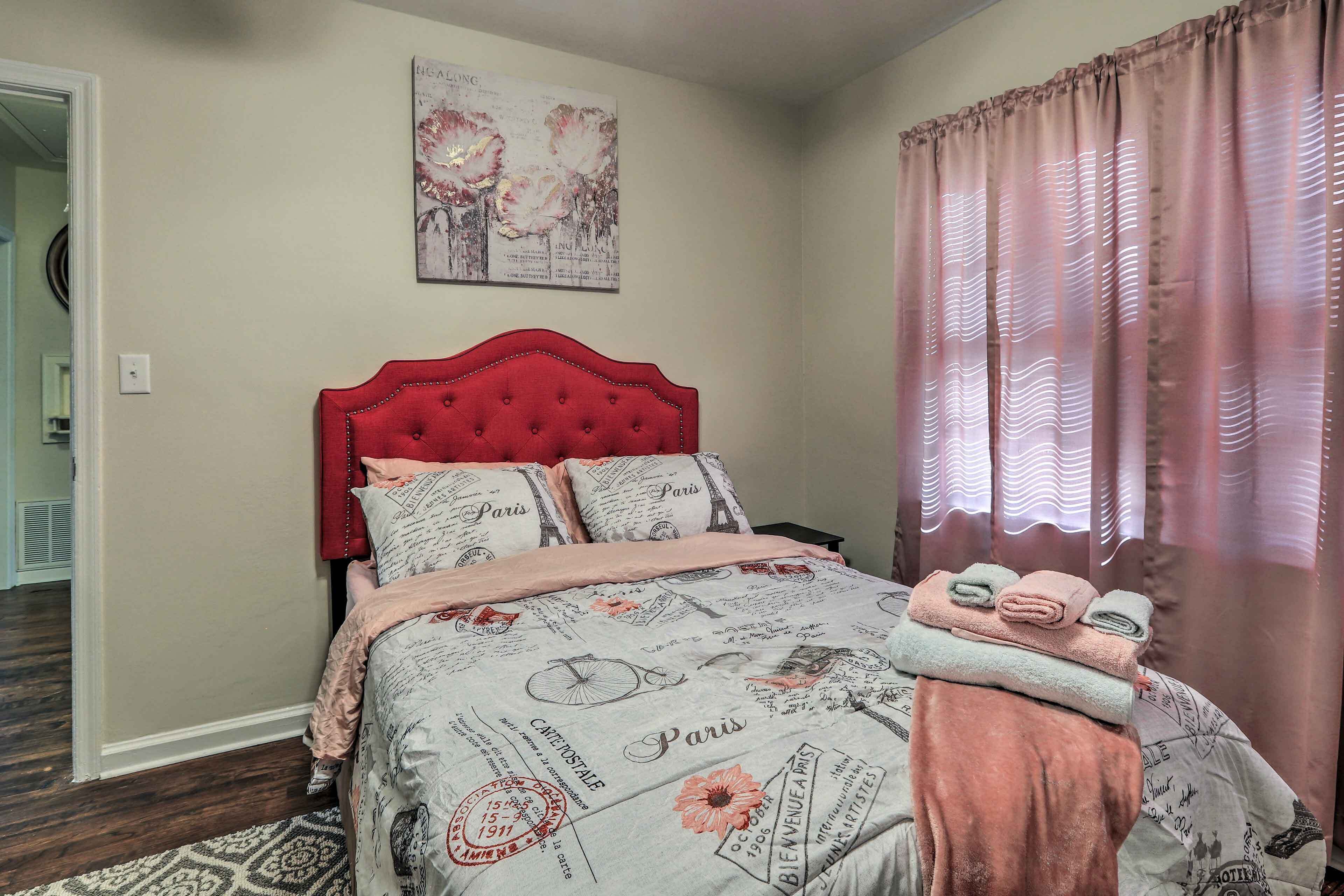 Un dormitorio con cabecera tapizada en rojo, cama con edredón con temática parisina y ventana con cortinas rosadas.