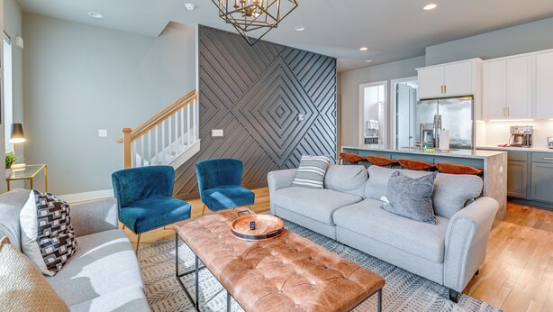 A modern living room with a grey sofa, blue armchair, and a brown ottoman. A geometric-patterned wall and a kitchen with white cabinets are visible. - Nashville Townhome w/ Rooftop Balcony & Game Room! (Nashville)