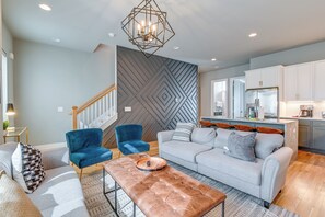 A modern living room with a grey sofa, blue armchair, and a brown ottoman. A geometric-patterned wall and a kitchen with white cabinets are visible. - Nashville Townhome w/ Rooftop Balcony & Game Room! (Nashville)