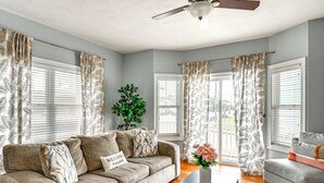 A living room with a beige sofa, a coffee table, and a grey armchair. There is a ceiling fan, a potted plant, and a window with curtains. - Inviting Norfolk Family Retreat: 1 Block to Beach! (Norfolk)