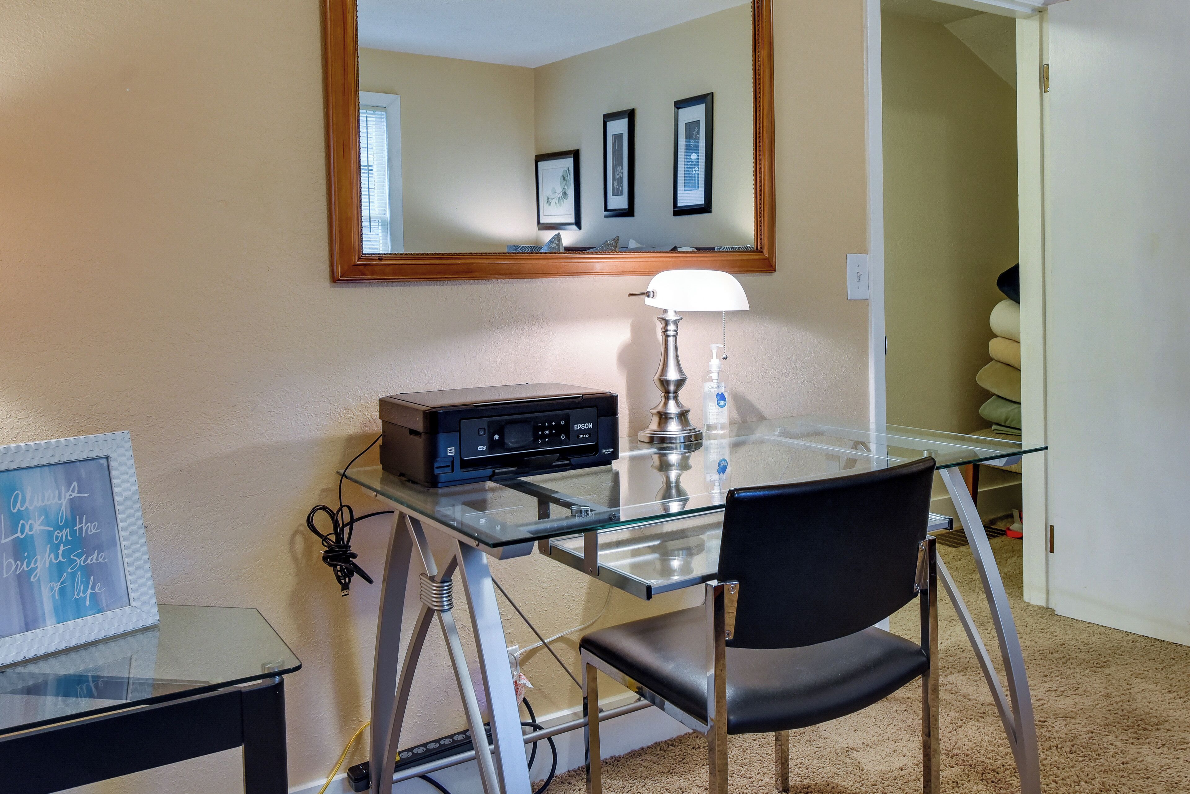 A room with a glass desk, a chair, a lamp, a printer, and a framed picture on the wall.