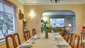 Une salle à manger avec une longue table dressée pour un repas, des chaises en bois, un vase de fleurs et une vue sur l’extérieur à travers une fenêtre munie de stores.