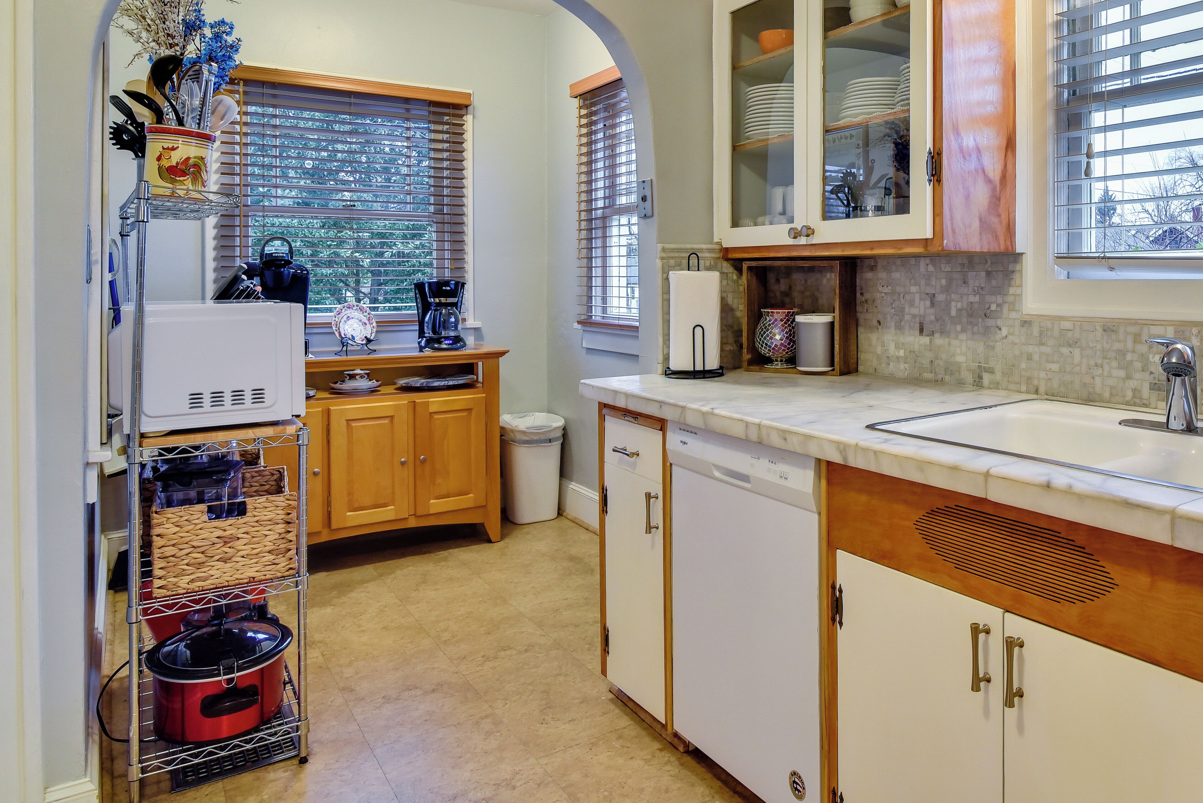 A kitchen with wooden cabinets, a dishwasher, a coffee maker, and a window with blinds.