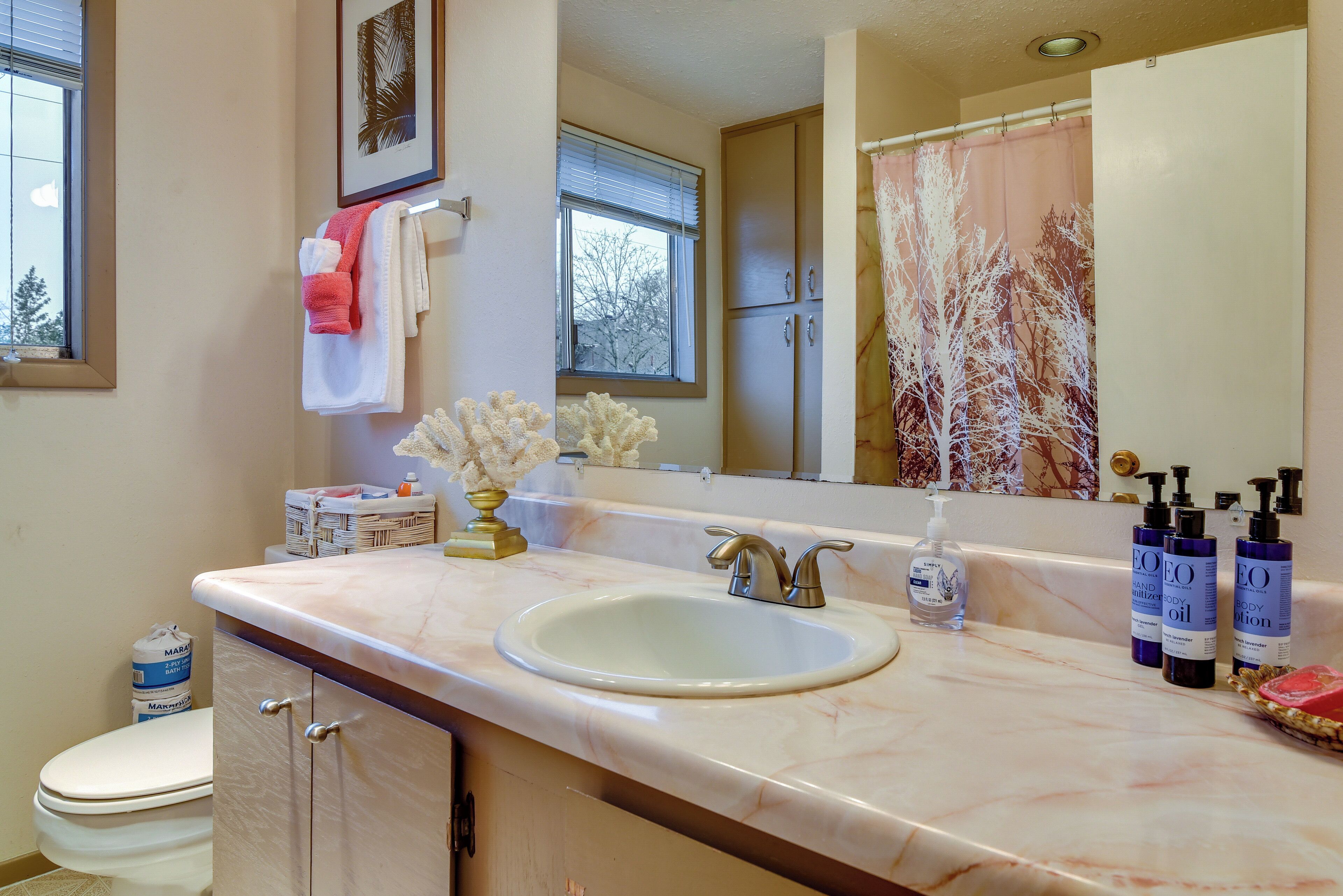 Une salle de bain avec un lavabo blanc, un meuble-lavabo et un grand miroir. On y trouve des articles de toilette sur le comptoir et une fenêtre avec des stores.