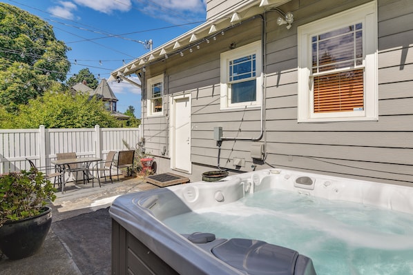 A hot tub in front of a house with a patio, a wooden fence, and a small table with chairs.