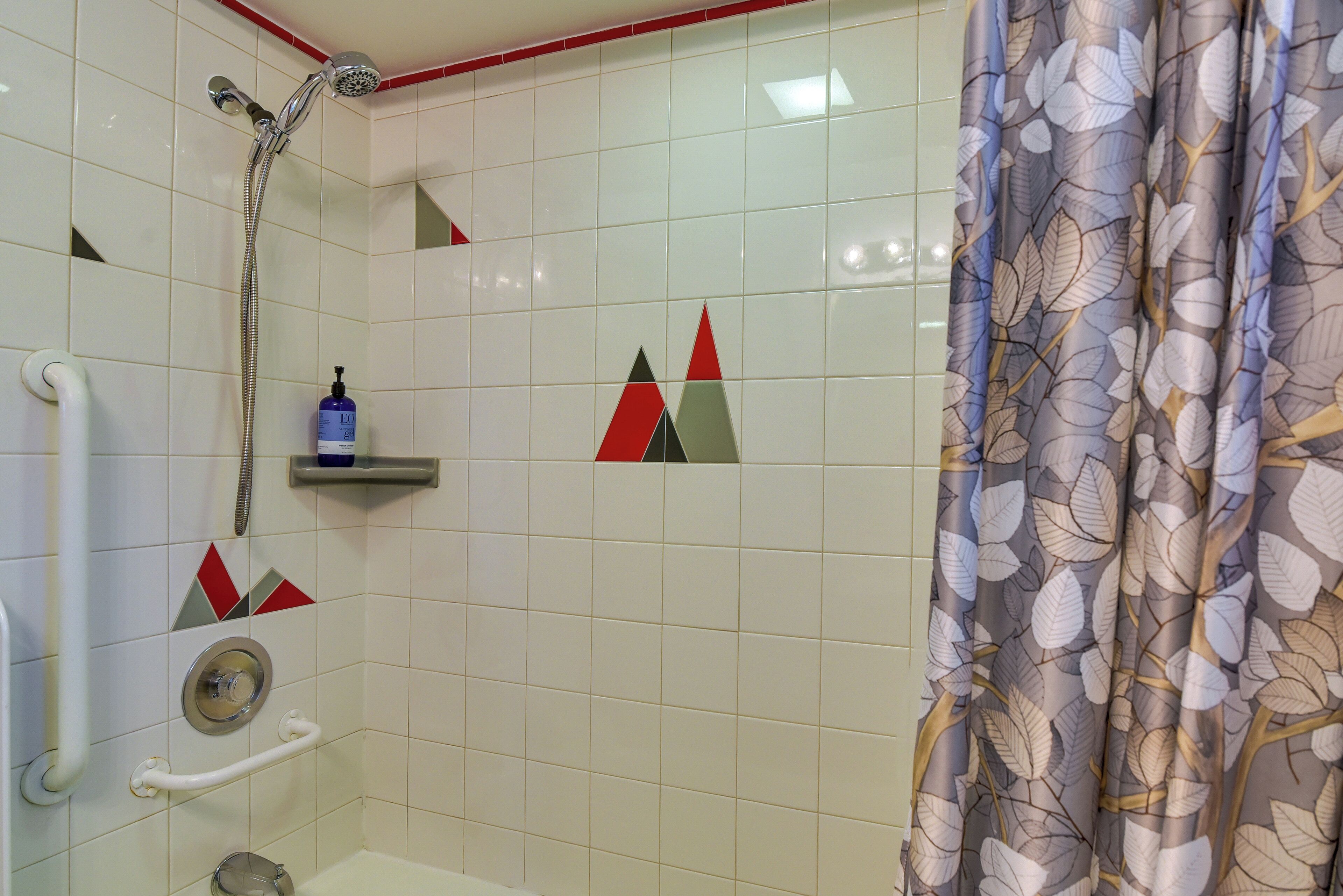 A bathroom with white tiles, a shower, a handrail, and a shelf with toiletries.