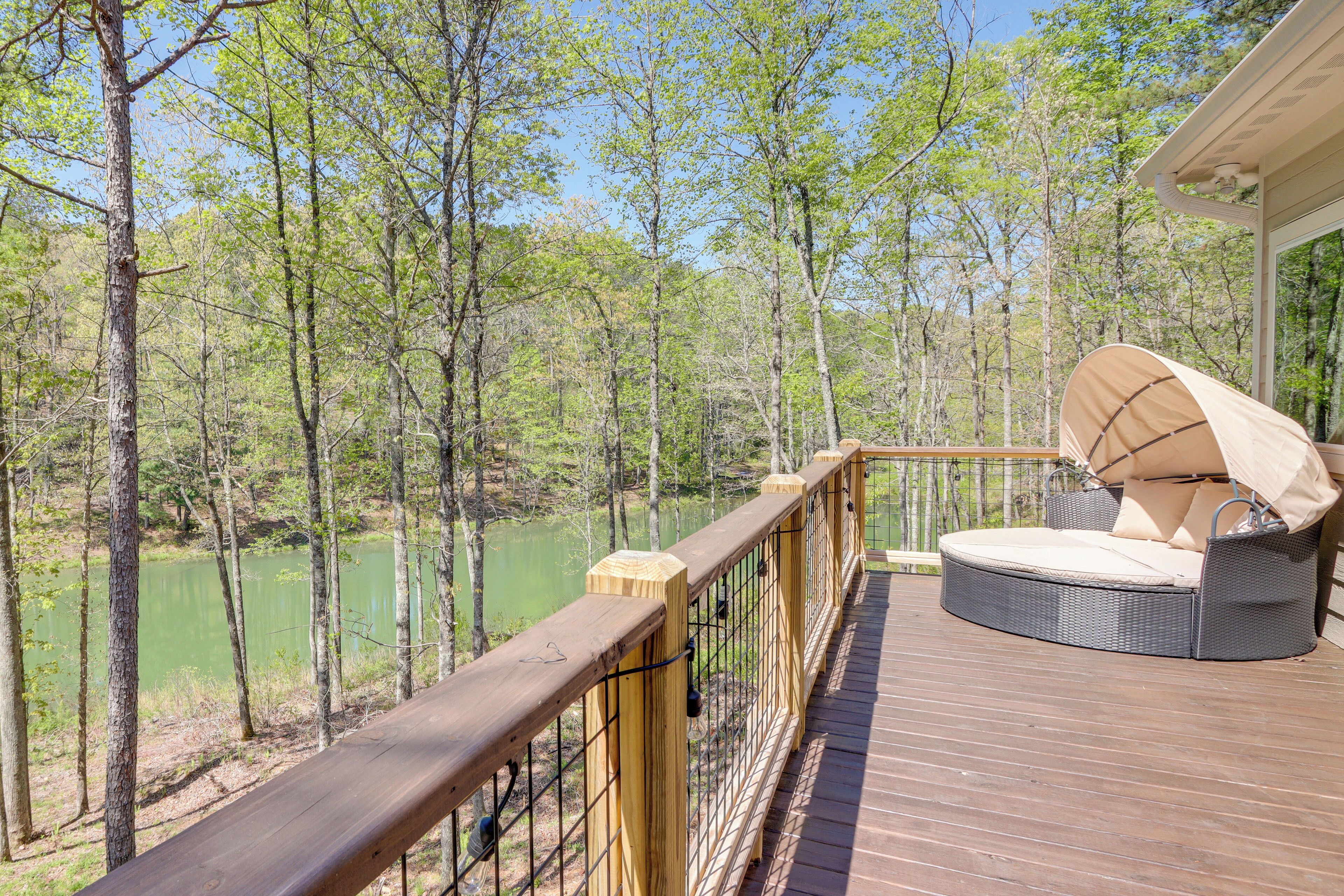 Une terrasse en bois avec un canapé rond en osier et des coussins, donnant sur une zone boisée et un plan d’eau.