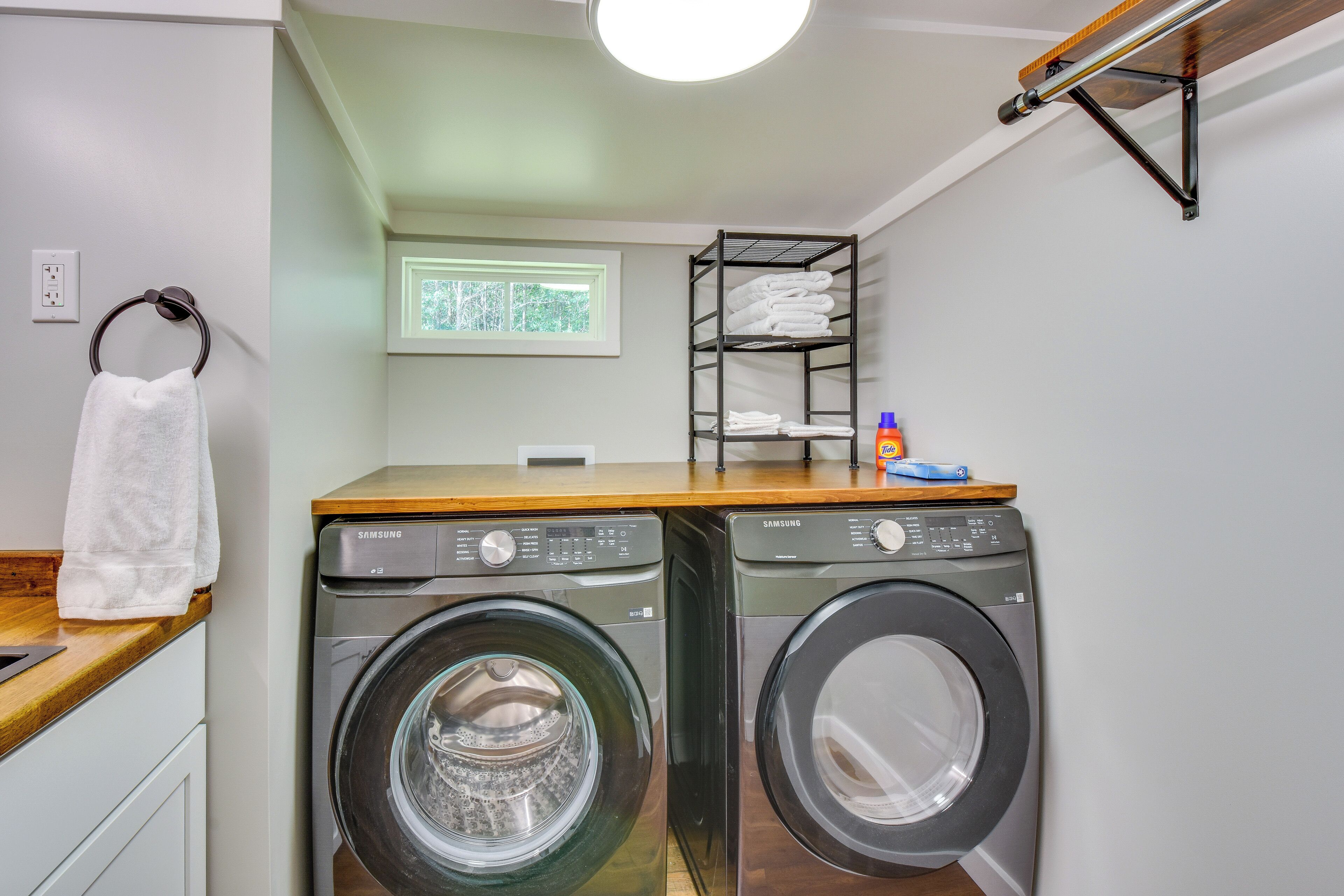 A laundry area with two front-loading washing machines, a wooden countertop, a towel rack, a shelf with folded towels, and a window with a view of trees.