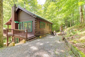 A log cabin with a porch, a gravel driveway, and a forested area.
