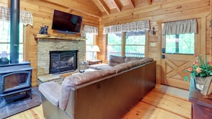 Ein gemütliches Blockhauss-Interieur mit Kamin aus Stein, einer Couch, einem Fernseher und einer Holzbalkendecke.