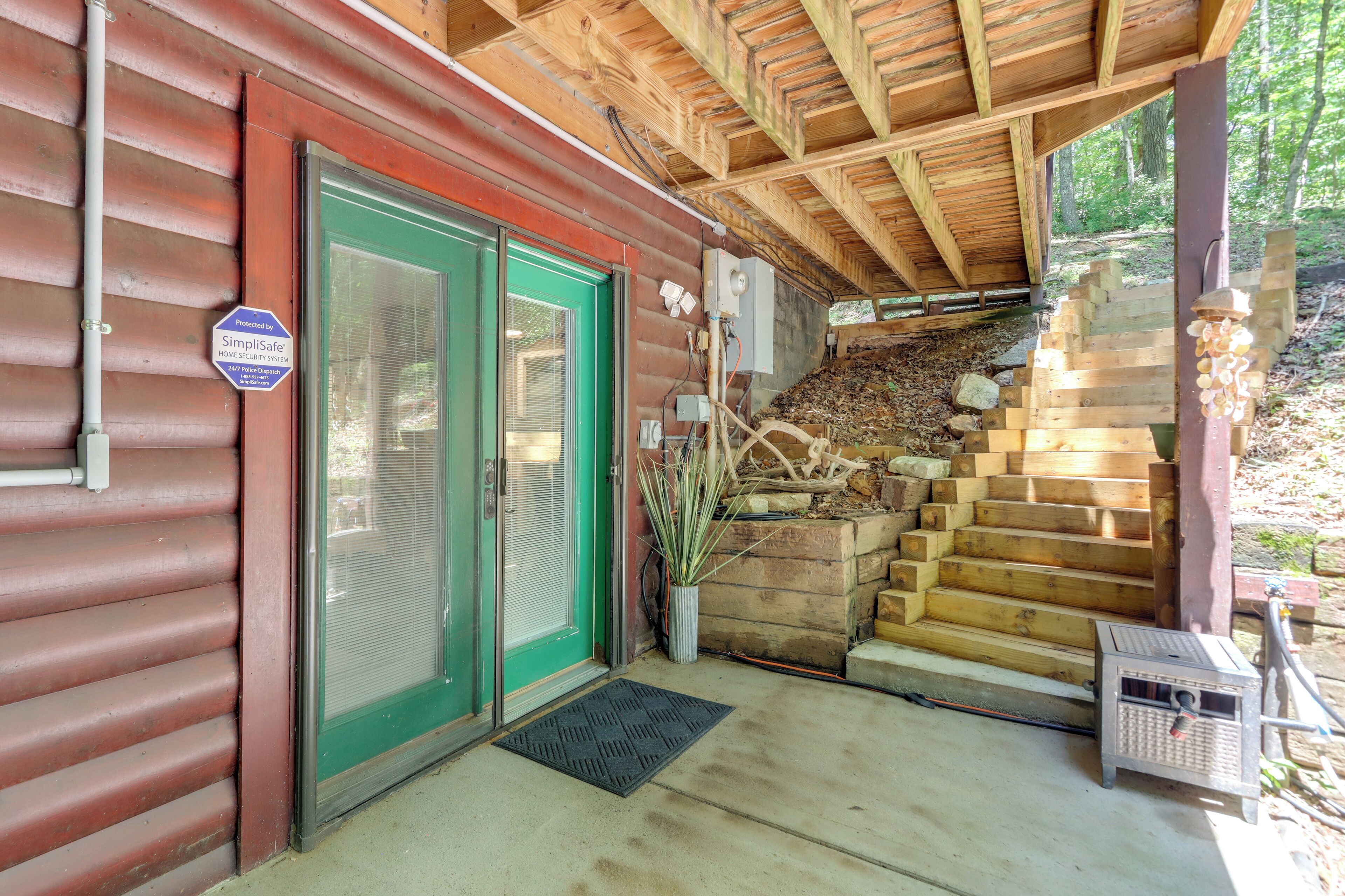 Une cabane en bois avec une porte verte, un panneau indiquant la présence d’un système de sécurité, un escalier menant à une terrasse en bois et un petit abri de jardin.