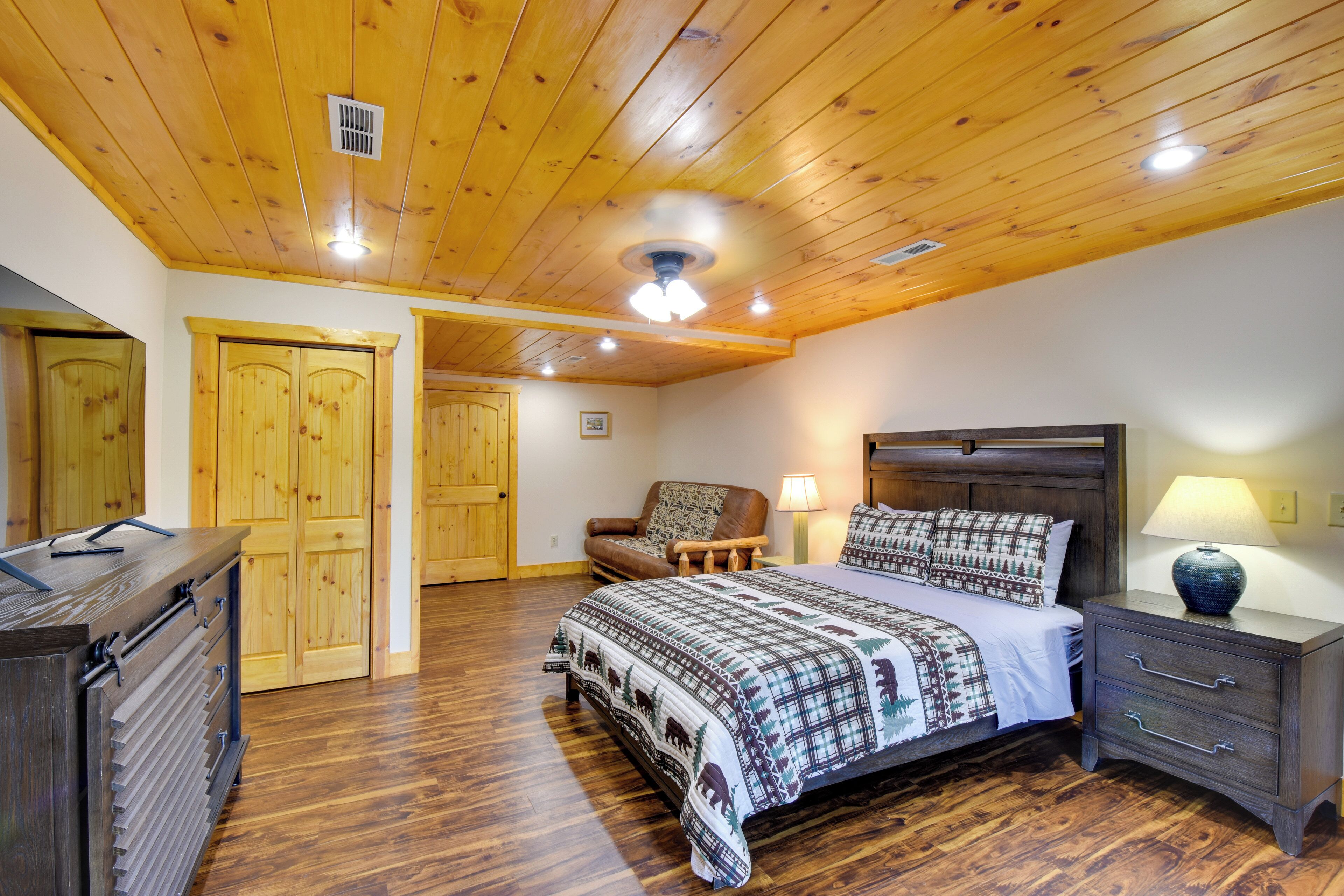 A bedroom with a wooden ceiling, a bed with a patterned bedspread, a nightstand with a lamp, a dresser, and a chair.