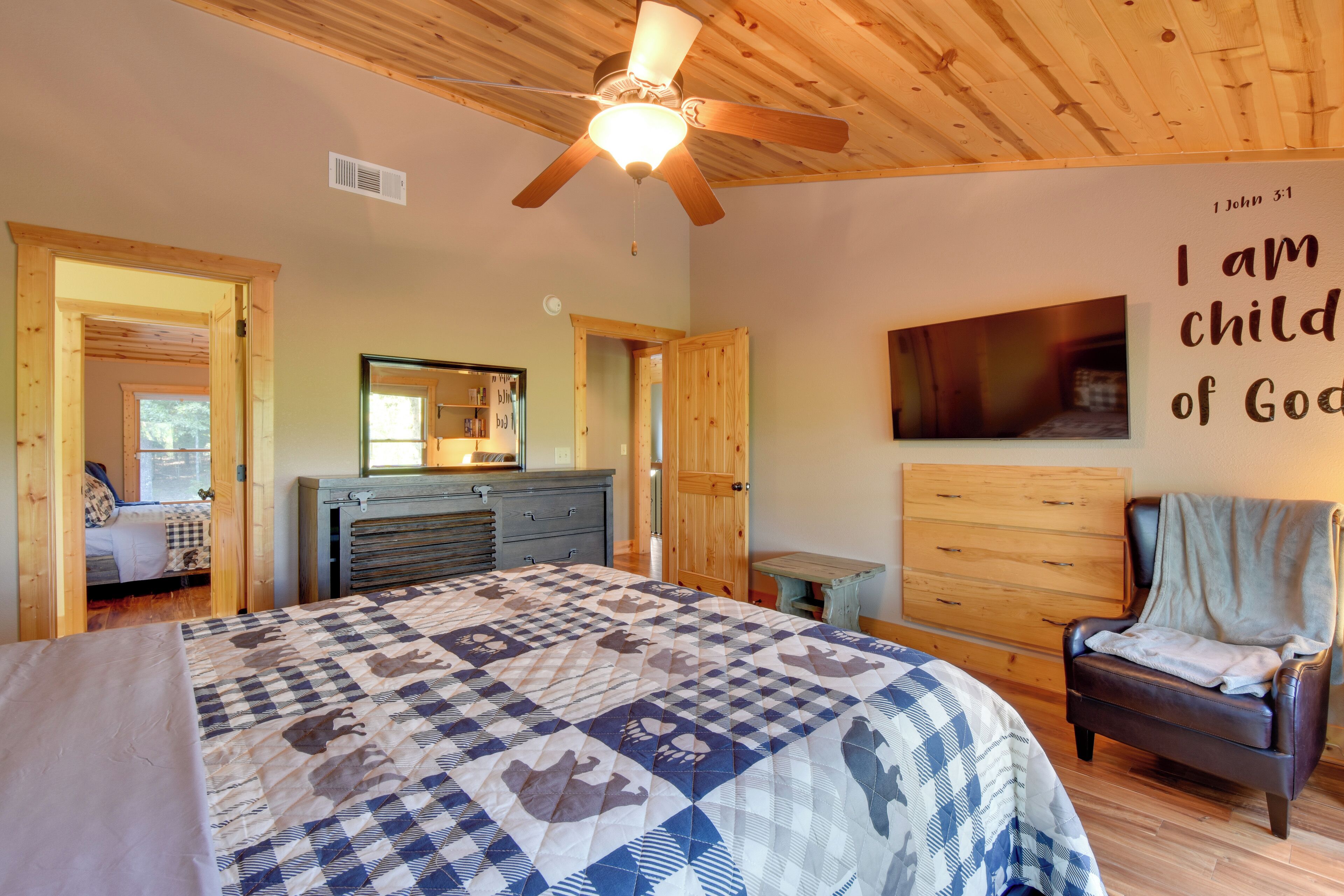 A bedroom with a large bed, a ceiling fan, a wooden dresser, a chair, and a wall with a religious quote.