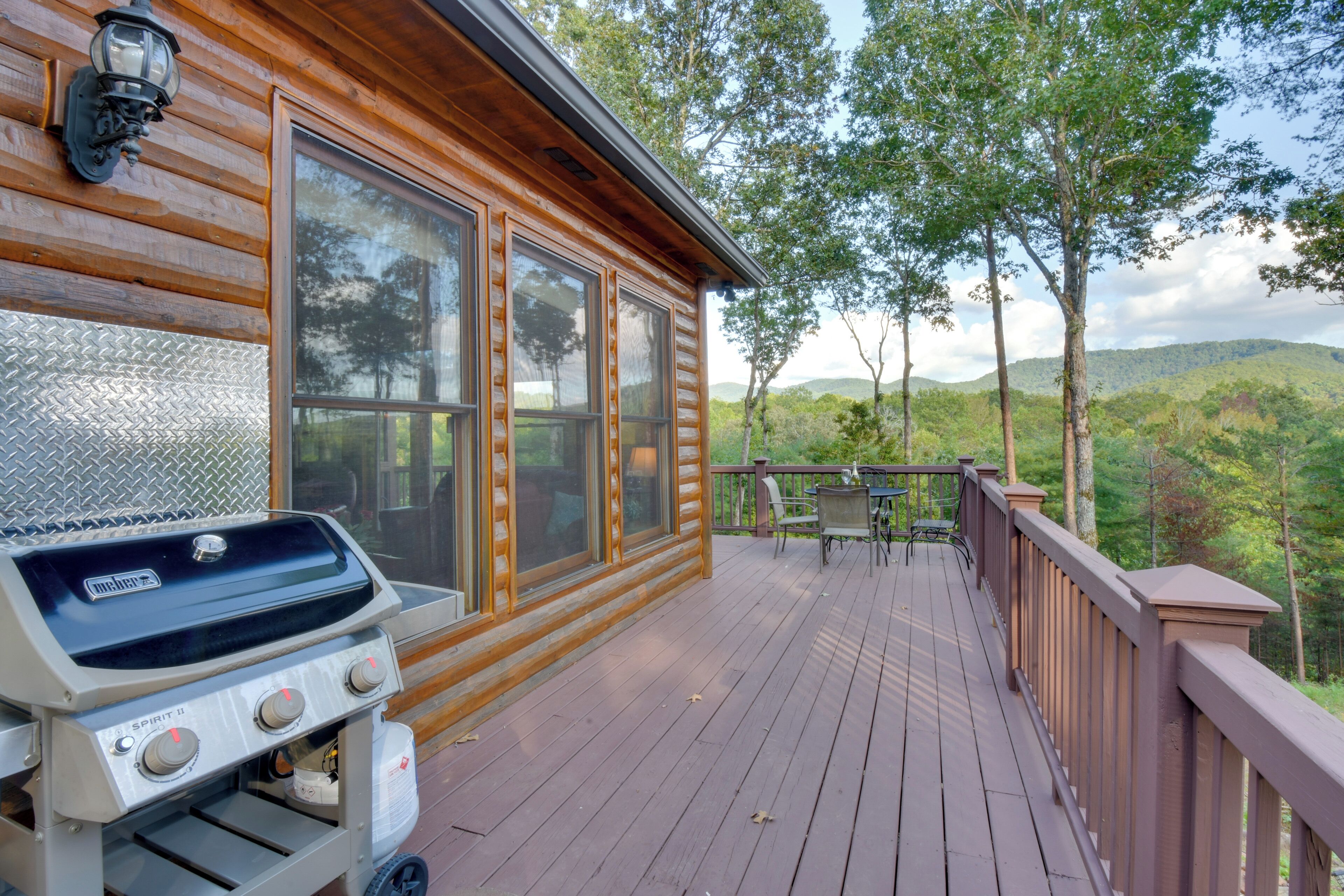 A wooden deck with a grill, a table, and chairs, overlooking a forested landscape.