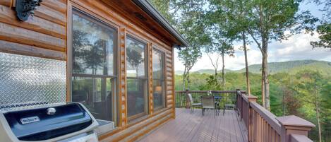 A wooden deck with a grill, a table, and chairs, overlooking a forested landscape.