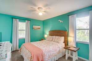 A bedroom with teal walls, a bed with patterned bedding, a ceiling fan, a bedside table with a lamp, and a window with curtains. - Coastal St Simons Retreat Near Gascoigne Bluff! (St. Simons Island)