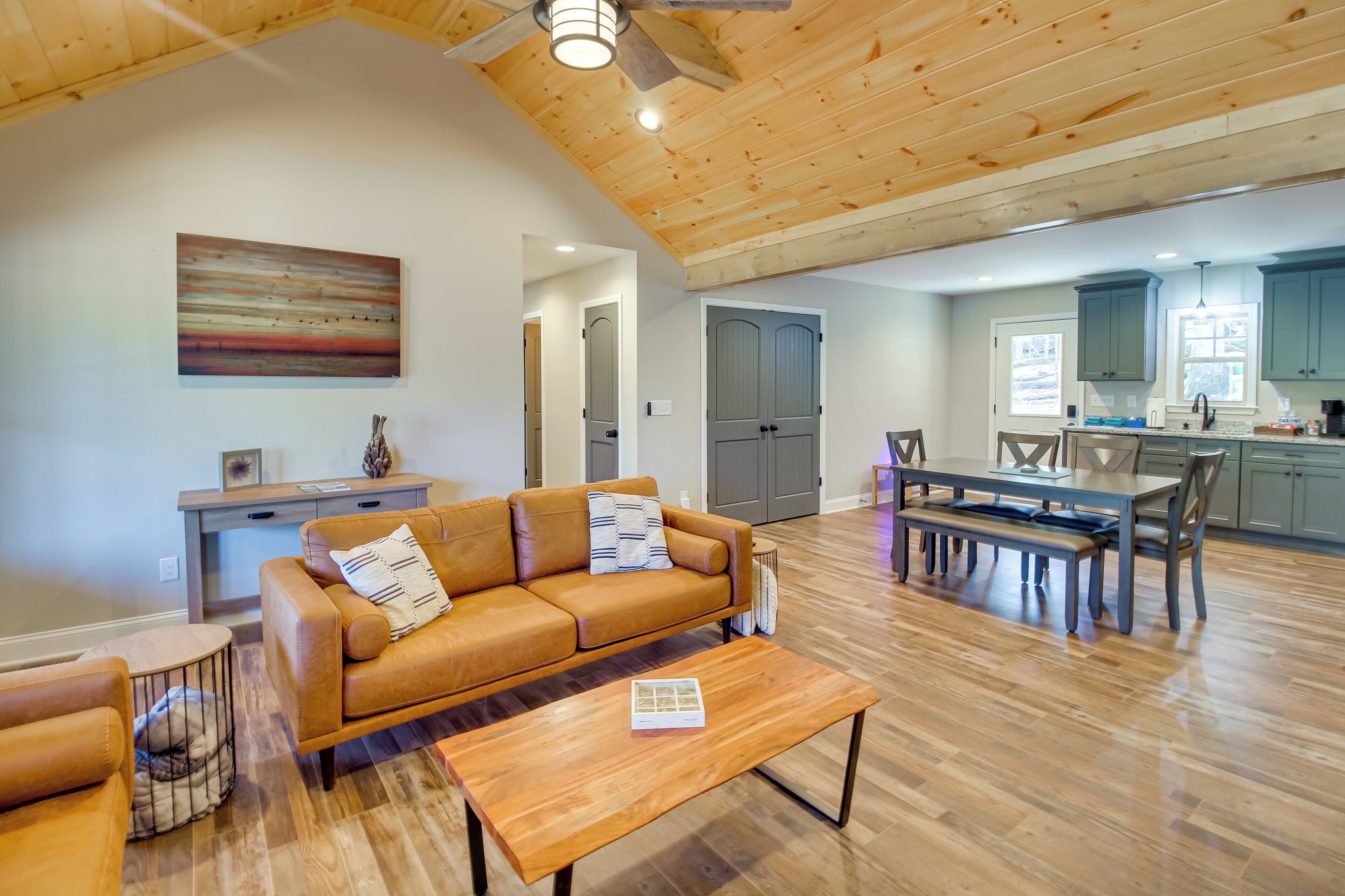 Un vaste salon doté d’un plafond en bois, d’un canapé en cuir marron, d’une table à manger avec des chaises, et d’un coin cuisine avec des placards et une fenêtre.