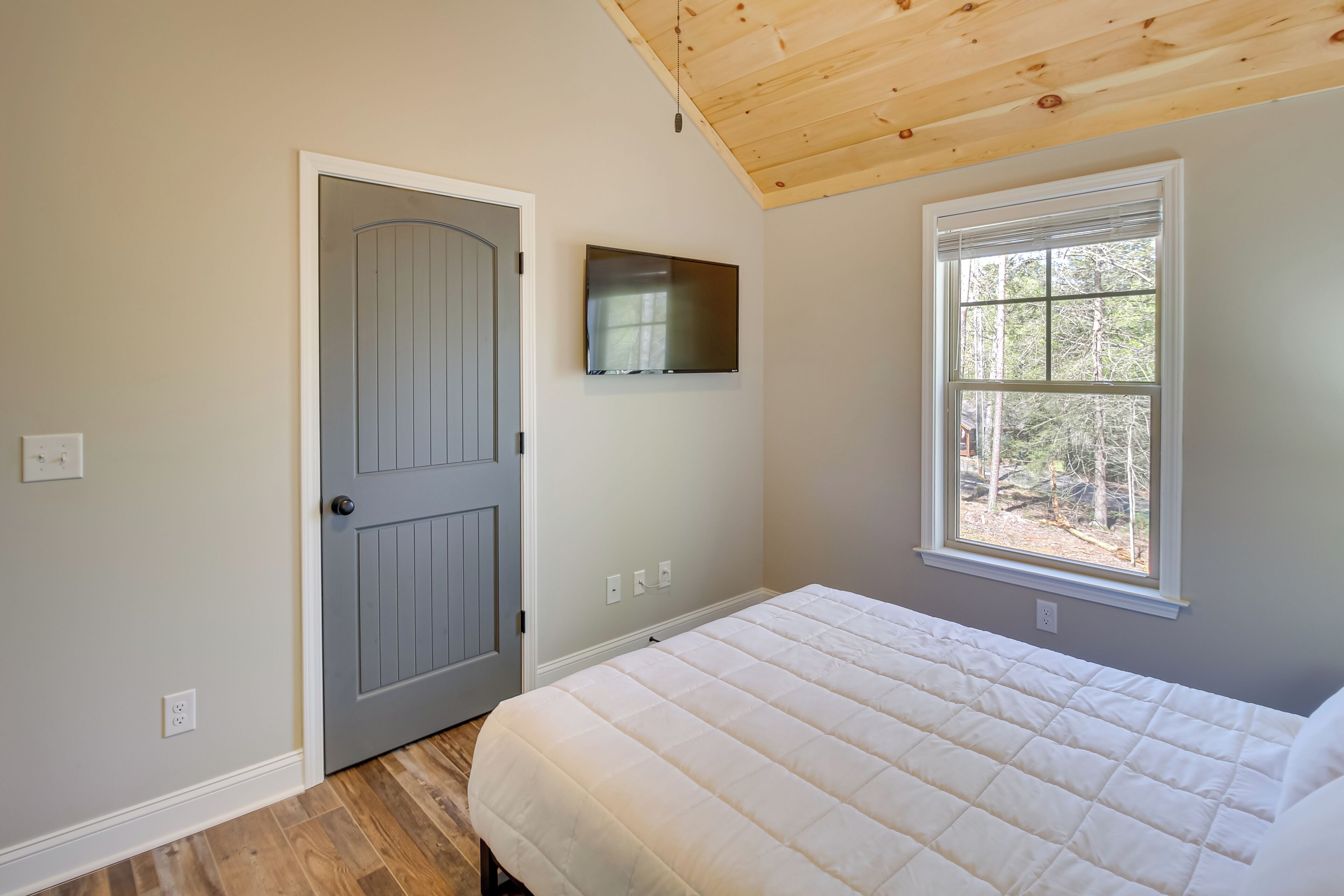 A bedroom with a bed, a door, a window, and a TV on the wall.