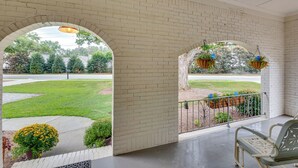 Une véranda couverte aux murs en briques, avec un banc, des jardinières suspendues et une vue sur une pelouse verdoyante et des arbres.