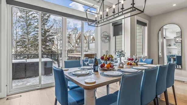 Un coin repas avec une grande table dressée pour six personnes, entourée de chaises bleues, et offrant une vue sur les arbres et un paysage enneigé grâce à de grandes fenêtres.