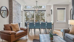 Un salon moderne doté d’une cheminée, de canapés en cuir, d’un coin repas avec des chaises et de grandes fenêtres offrant une vue sur les arbres.