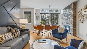 A modern living room with a grey sofa, blue armchairs, a dining area with orange chairs, a fireplace, and a large window offering a view of trees. - NEW! Oberhofen by SSR Top View HOT TUB 5BR ( BL# RES-11737 ) (Canmore)