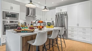 A modern kitchen with white cabinetry, stainless steel appliances, a central island with bar stools, and pendant lighting. - NEW! Oberhofen by SSR Top View HOT TUB 5BR ( BL# RES-11737 ) (Canmore)