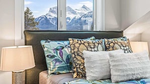 A bedroom with a bed, pillows, a lamp, and a window with a mountain view. - NEW! Oberhofen by SSR Top View HOT TUB 5BR ( BL# RES-11737 ) (Canmore)