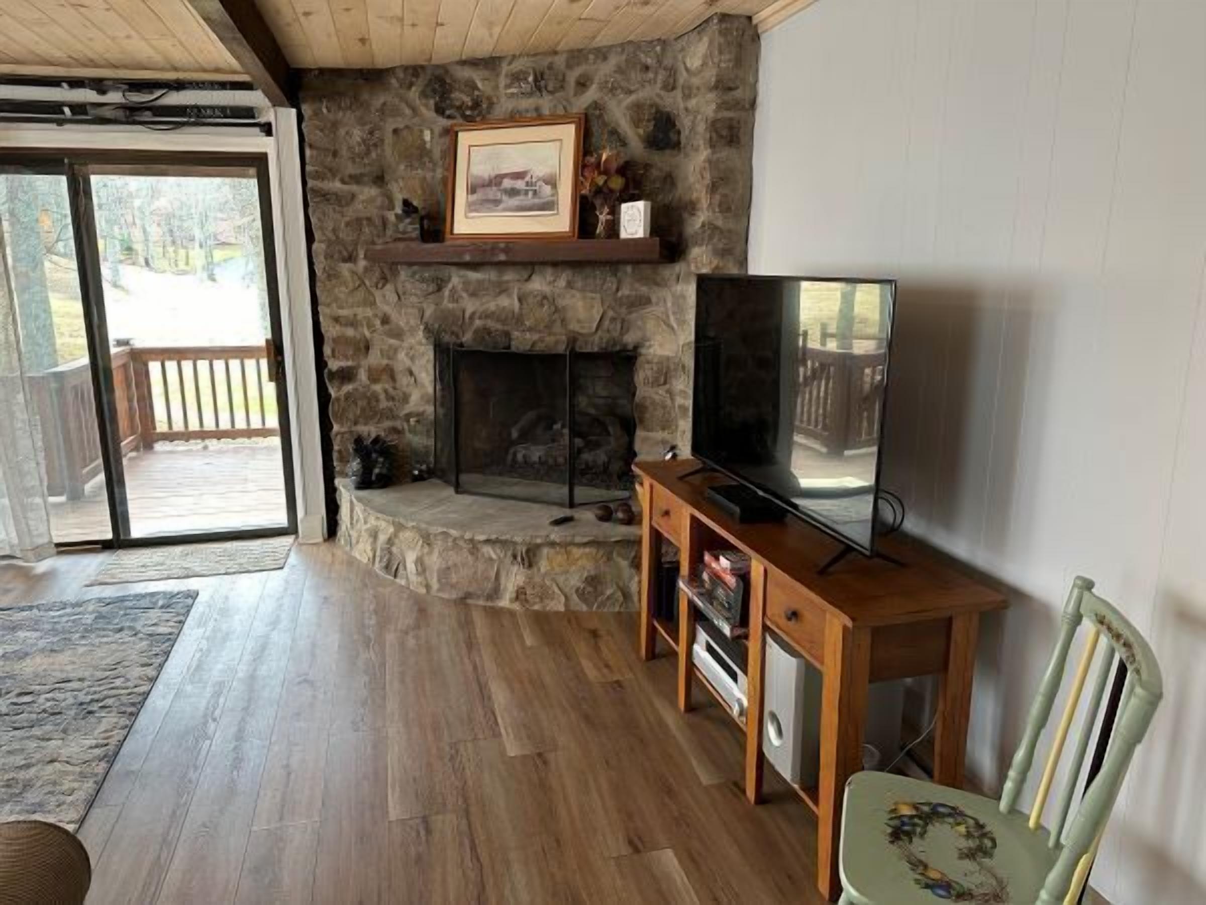 A living room with a stone fireplace, a television on a wooden stand, and a view of the outdoors through a glass door.