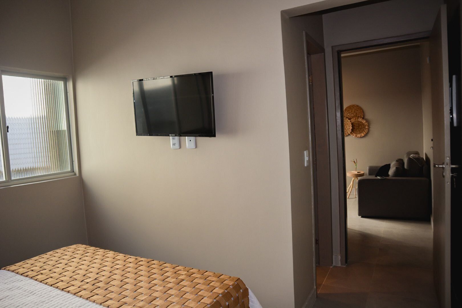 A hotel room with a bed, a television mounted on the wall, and a doorway leading to a living area with a sofa and a decorative wall hanging.