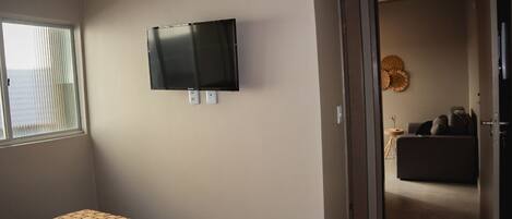 A hotel room with a bed, a television mounted on the wall, and a doorway leading to a living area with a sofa and a decorative wall hanging.