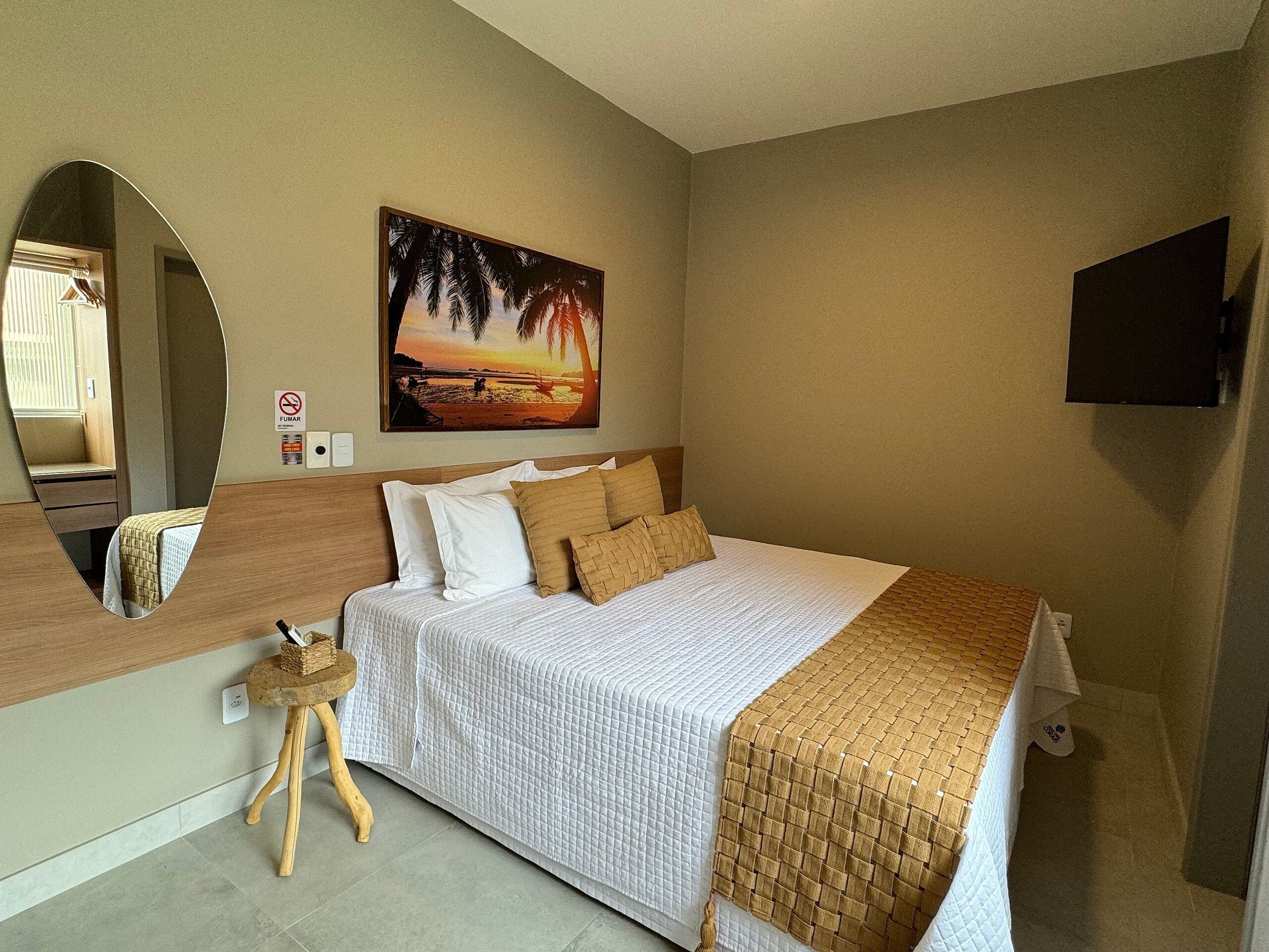 Une chambre à coucher avec un lit, un miroir, un tableau représentant un coucher de soleil, une télévision et un petit tabouret.