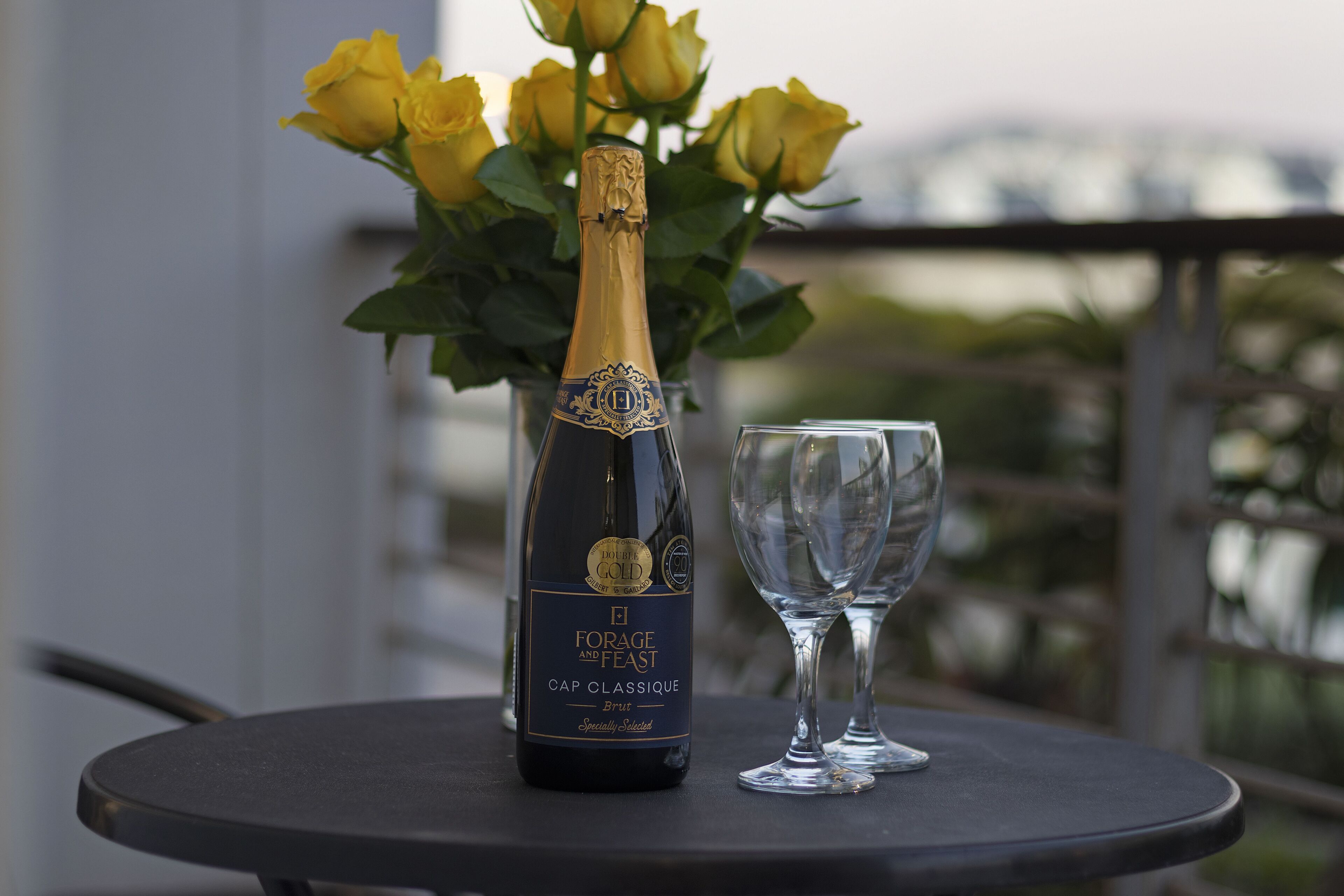 Een fles Forage and Feast Cap Classique Brut champagne op een tafel, samen met een glas en gele rozen.