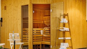A sauna room with wooden walls, benches, and a glass door leading to a shower area. There are towels and a bottle on a table. - Seepark Hotel am Wandlitzsee (Wandlitz)