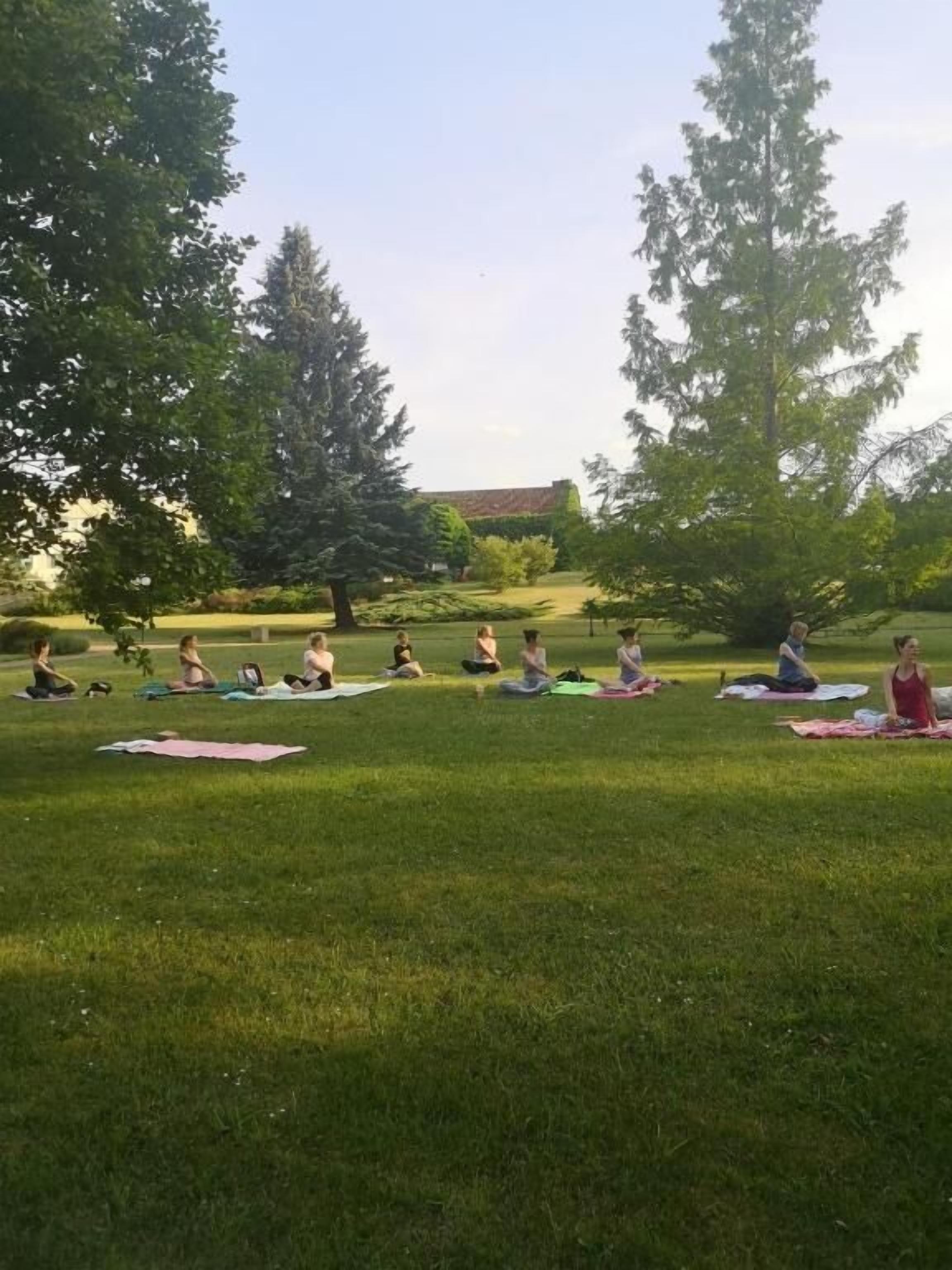 A group of people sitting on mats in a grassy outdoor area with trees and a building in the background.