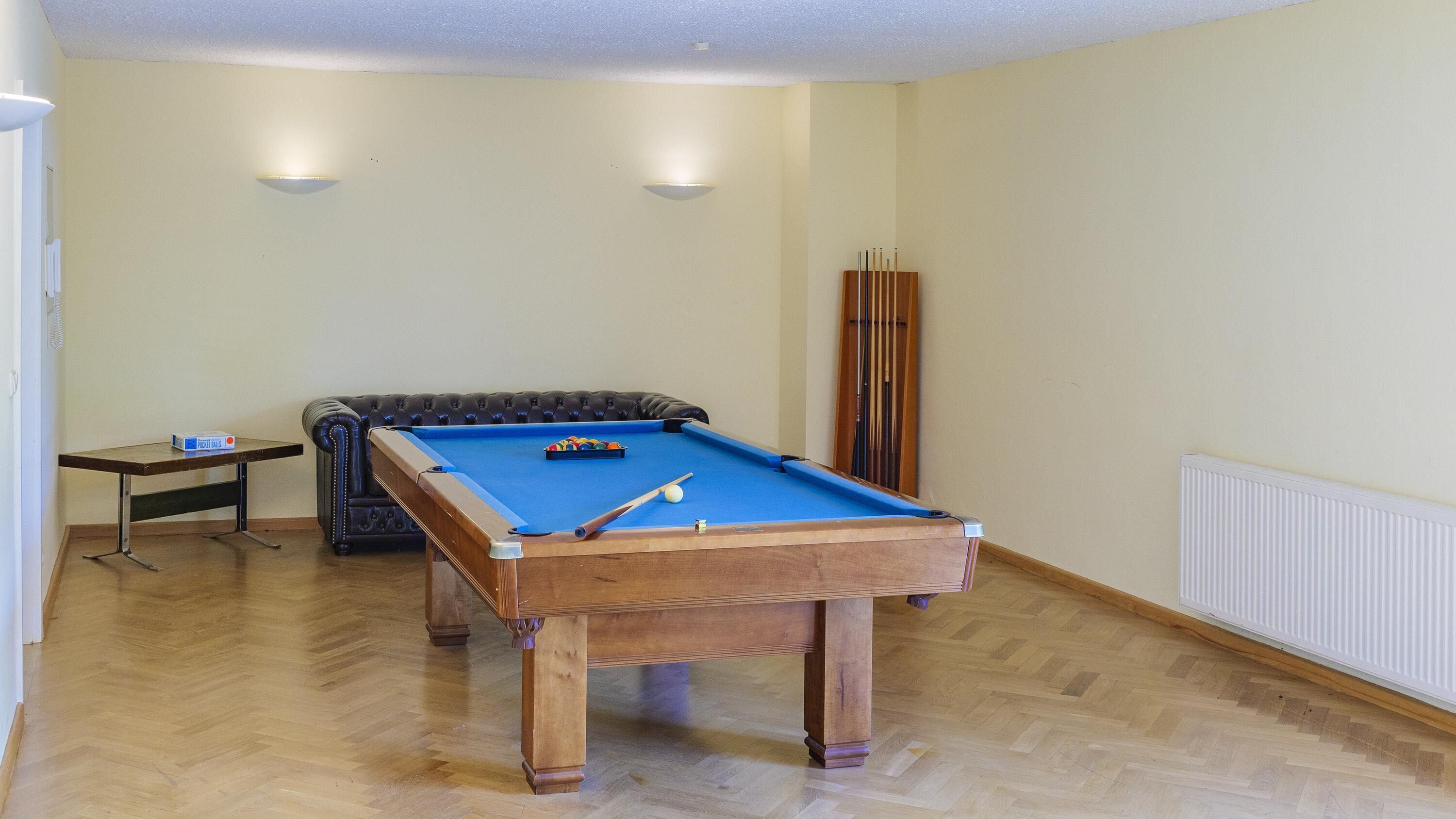 Una mesa de billar con superficie de fieltro azul, una pequeña mesa con una caja de bolas de billar y un sofá de cuero en una habitación con suelo de madera y paredes blancas.