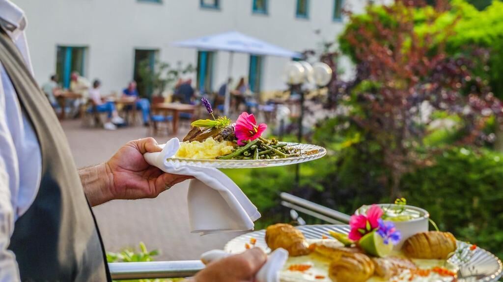 A person holding a plate with food and flowers, with a dining area and outdoor seating in the background.